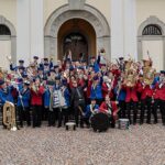 Eine Gruppe von Menschen in blauen und roten Uniformen mit verschiedenen Instrumenten. Am Rand hält jeweils eine Person einen Fahnen.