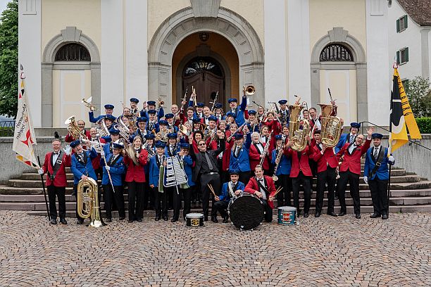 Eine Gruppe von Menschen in blauen und roten Uniformen mit verschiedenen Instrumenten. Am Rand hält jeweils eine Person einen Fahnen.